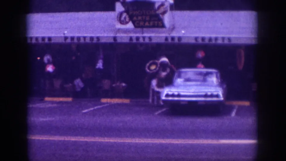 Home movie frame c.1963, Memphis Tennessee, costumed street figure with distinctive circular props beside a pale American sedan outside an arts and crafts retailer.