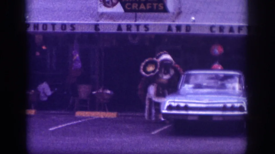 8mm home movie, Memphis Tennessee c.1963, costumed street figure with circular props beside a classic American sedan, commercial storefront signage in background.