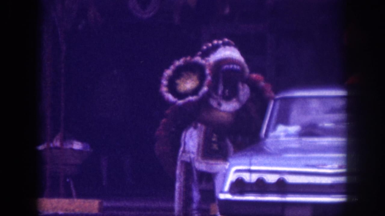Person in large double feathered headdress costume standing closer beside a light-colored classic American sedan, heavy purple color cast and grain.