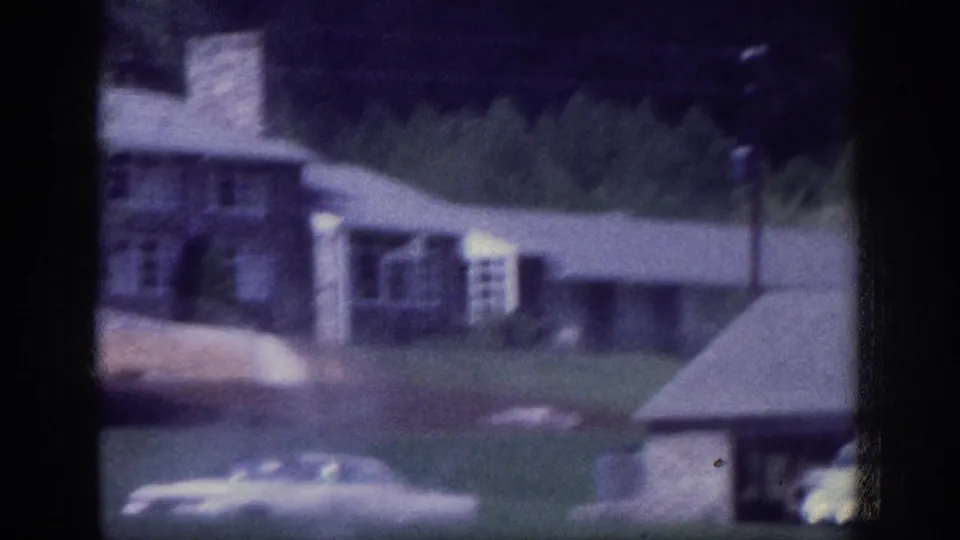 8mm home movie circa 1963, possibly Memphis Tennessee suburb: stone-facade residential house with parked cars, green lawn, and mature trees, mild residual purple film cast.
