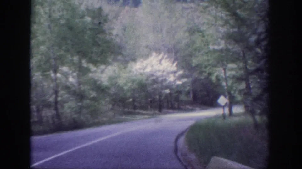 8mm home movie, circa 1963, Memphis Tennessee: winding forested two-lane road with a white diamond warning sign viewed through a vehicle windshield.