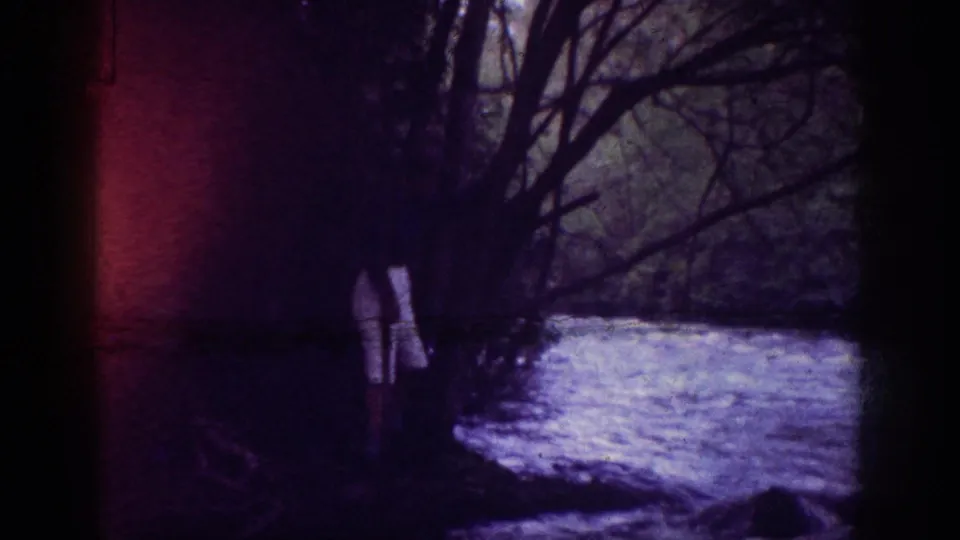 Adult figure in light-colored clothing near a river or creek bank framed by bare branches, with a red light-leak at frame left; 8mm home movie, 1963.