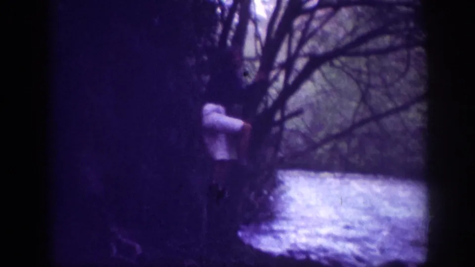 Adult figure in white or light top standing at a wooded riverbank framed by tree trunks and bare branches; heavy indigo cast on 8mm home movie stock, 1963.