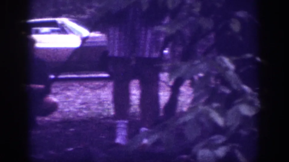 Era-appropriate light-colored sedan and wooden fence or covered structure in a wooded yard; 8mm home movie footage consistent with Memphis Tennessee circa 1963.