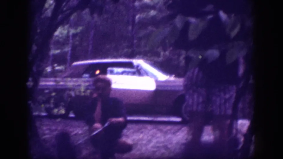 Mid-sequence 8mm frame from a 1963 Memphis, Tennessee roadside stop — large American sedan at center-right, adult figure crouching at lower left against a dense wooded backdrop, violet film cast.