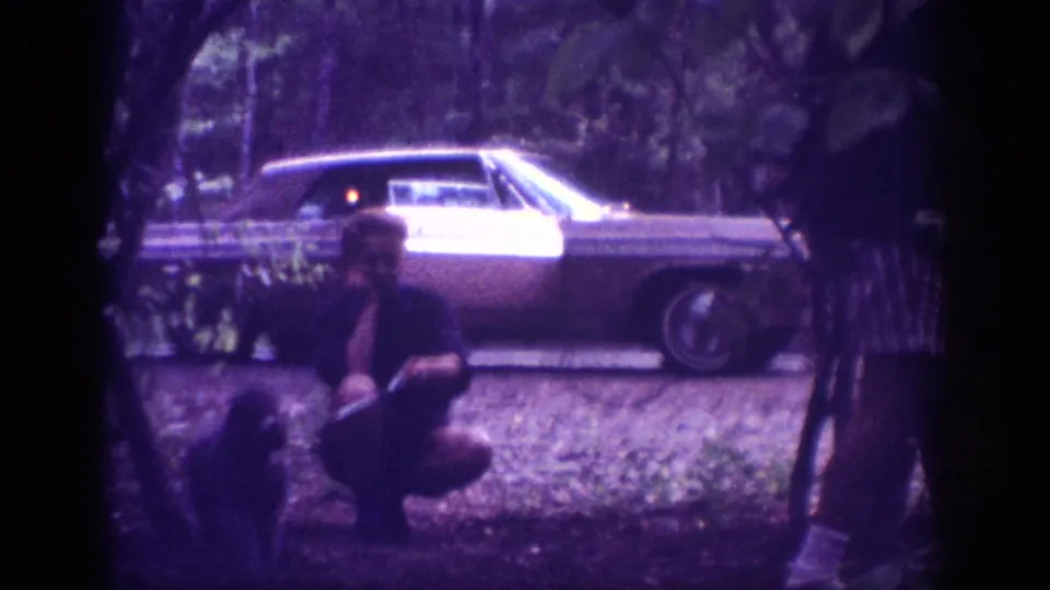 Final frame of a 1963 Tennessee roadside sequence — adult in dark clothing crouches near the rear of a large American sedan, dense woods behind, aged film violet color cast and visible grain throughout.