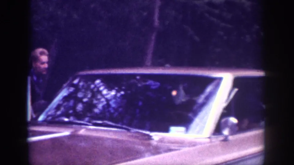 Wide, flat hood of a large early-1960s American sedan fills the frame; a woman stands near the vehicle in a forested roadside setting in this 8mm home movie.