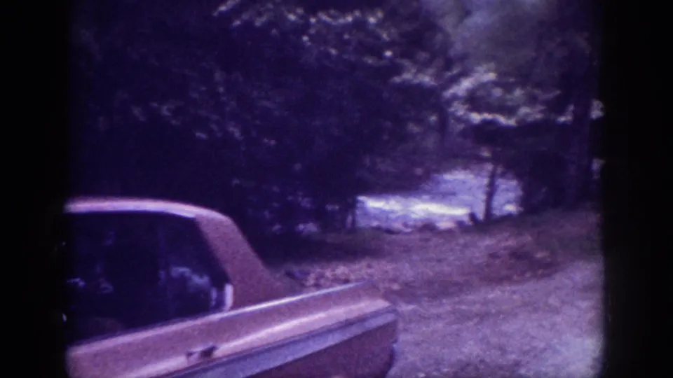Rear quarter of a large early-1960s American sedan on a wooded dirt road; trees crowd both sides and a lighter area suggesting water or a clearing is visible in mid-ground, 8mm footage.
