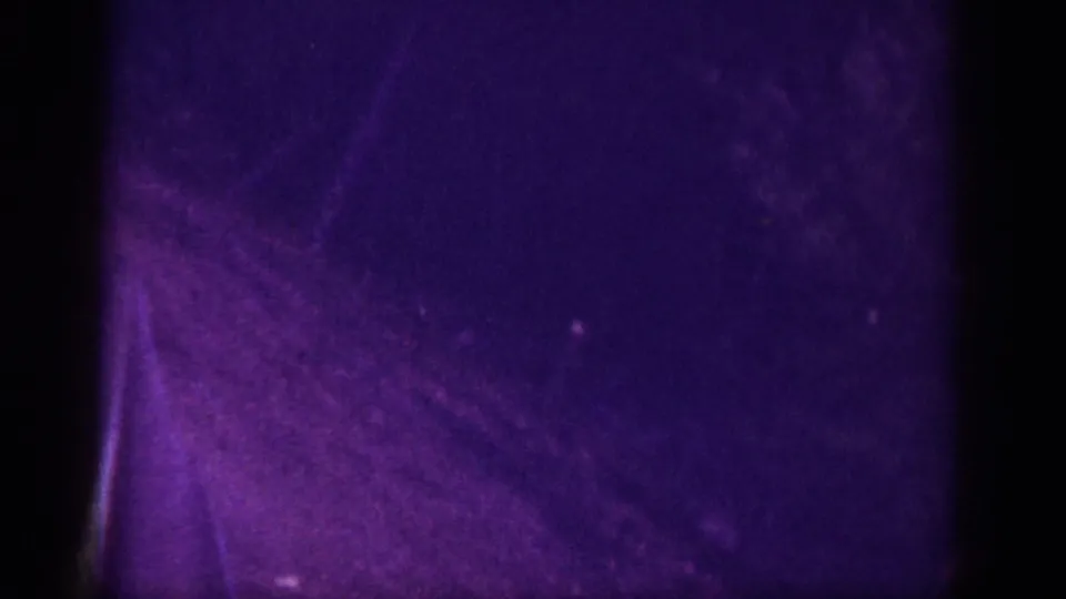 Severely underexposed frame from a 1963 8mm driving clip; purple-violet cast and dense grain obscure nearly all detail, with faint road or terrain texture.