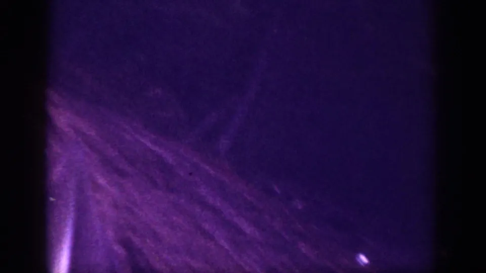 Dark 8mm frame from a 1963 road-trip clip; faint curving road visible through heavy grain and purple-violet dye shift, with a light artifact at lower frame edge.