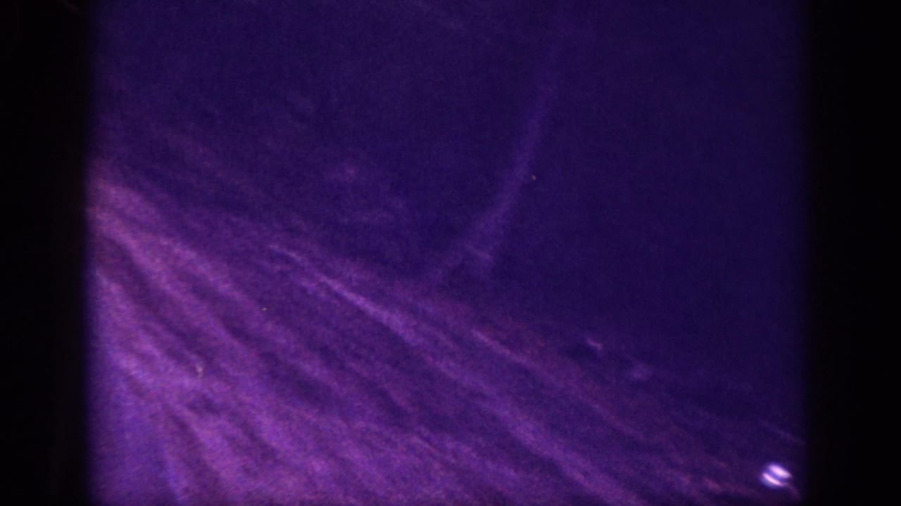 Abstract purple-cast 8mm frame showing diagonal scratches and motion blur over dark ground, with a small bright point of light.