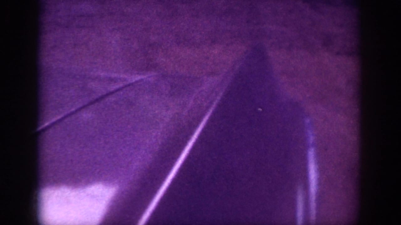 Mid-clip 8mm frame, c.1963, Memphis area — pale blurred mass at lower-left may indicate rear of departing car, framed by diagonal scratches and deep purple cast.