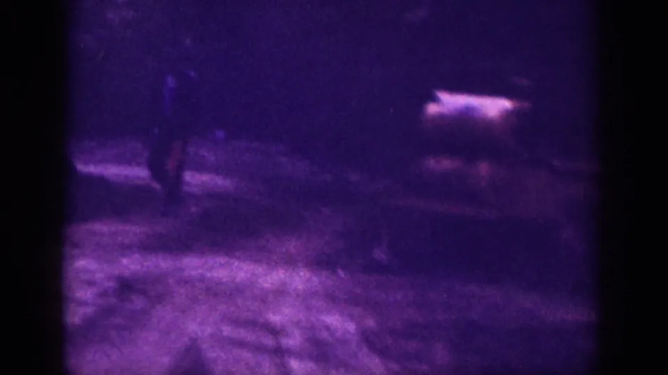 Clearest frame in a 1963 8mm home movie from Memphis, Tennessee; a dark male silhouette stands near an unidentified object on an unpaved surface under purple-shifted film color.