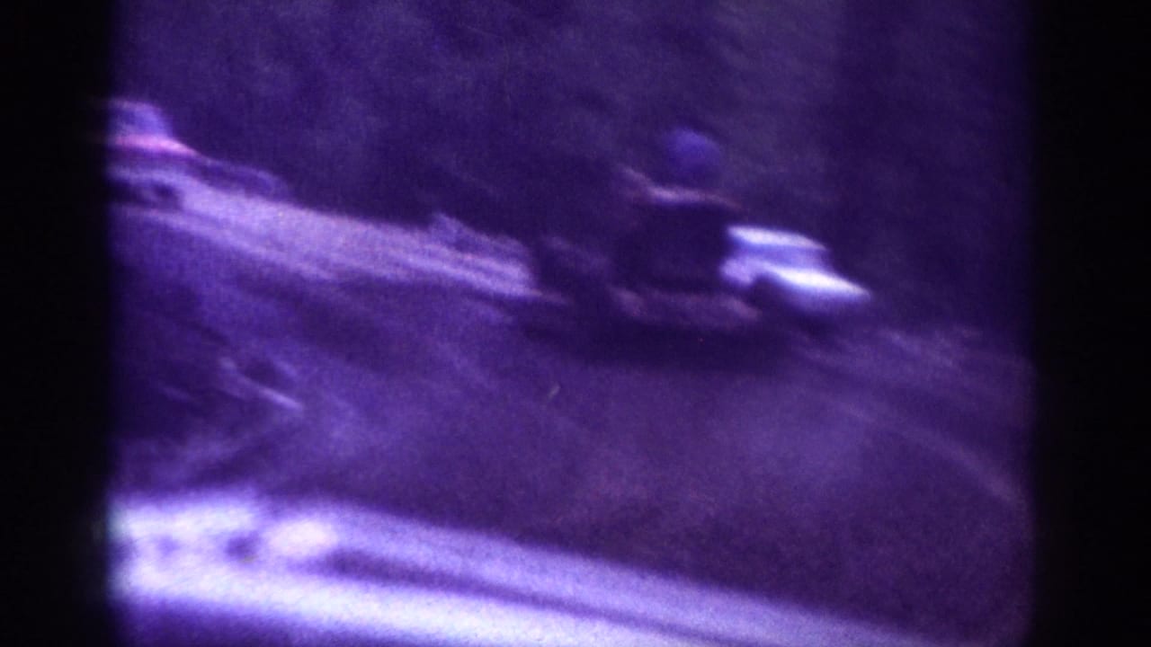 Blurred 8mm frame of vehicles on a road, purple color cast and motion blur; detail remains limited.