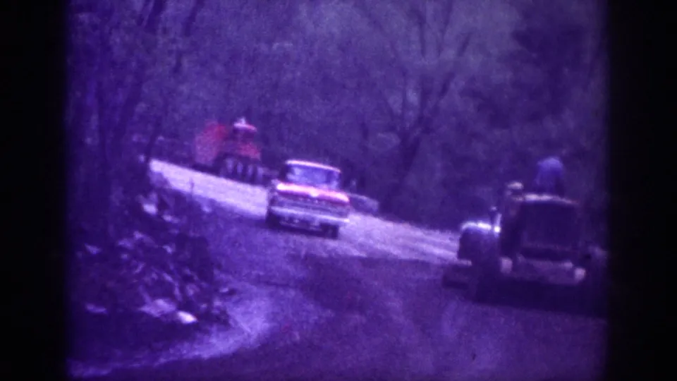 8mm home movie frame circa 1963 showing a winding mountain road with a pickup truck, a second vehicle, bare hillside trees, a rocky embankment, and a figure near the truck bed.