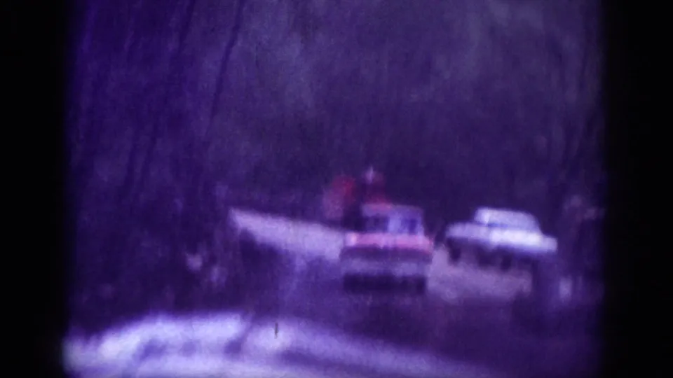 8mm home movie frame circa 1963 showing a pickup truck and a light-colored sedan on a narrow winding mountain road lined with bare trees.