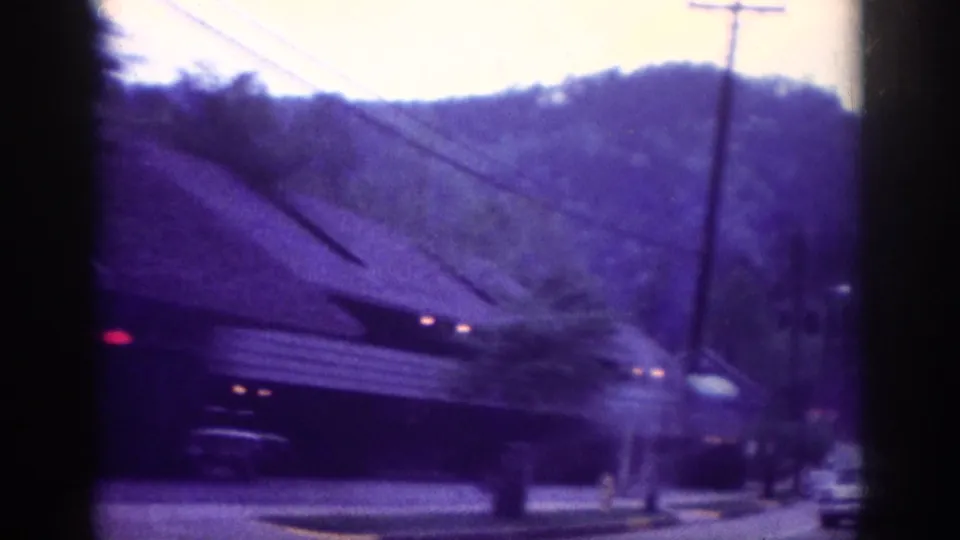 1963 8mm home movie: roadside building with low-pitched roof, utility poles, red vehicle tail lights, and a wooded mountain hillside — mid-century American roadside stop.