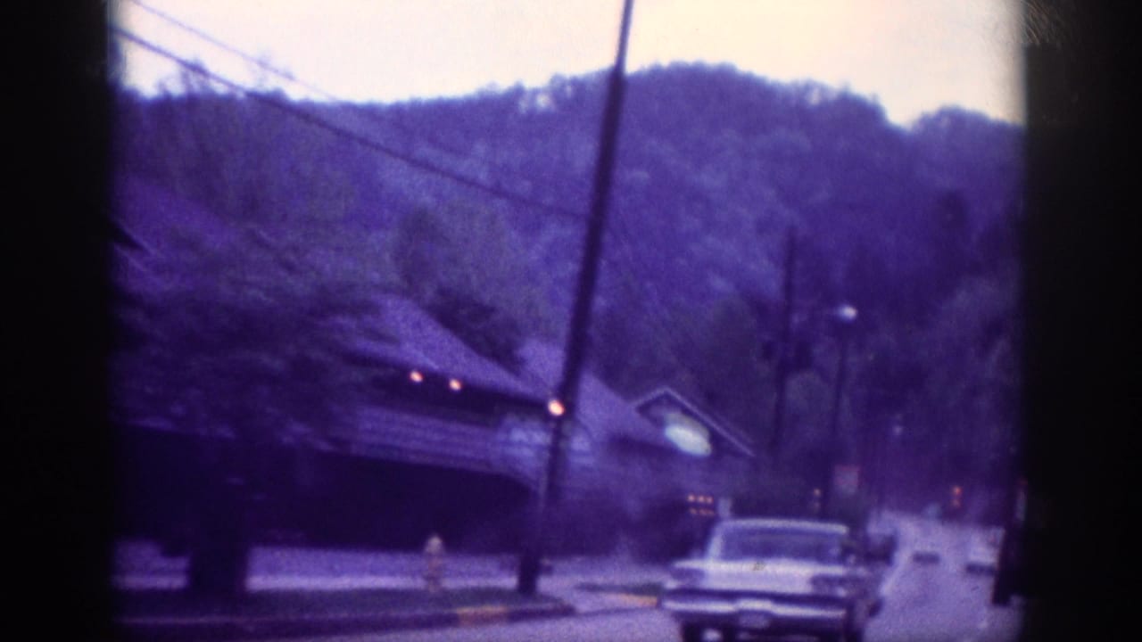 Two-lane road with an early 1960s American sedan in the foreground, utility poles along the left, and a tree-covered ridge beyond roadside buildings.