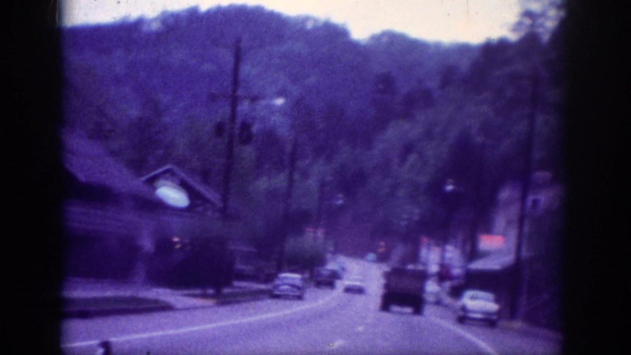 Passenger-perspective 8mm footage of several 1960s cars driving along a Tennessee roadside commercial strip backed by wooded hills, circa 1963.