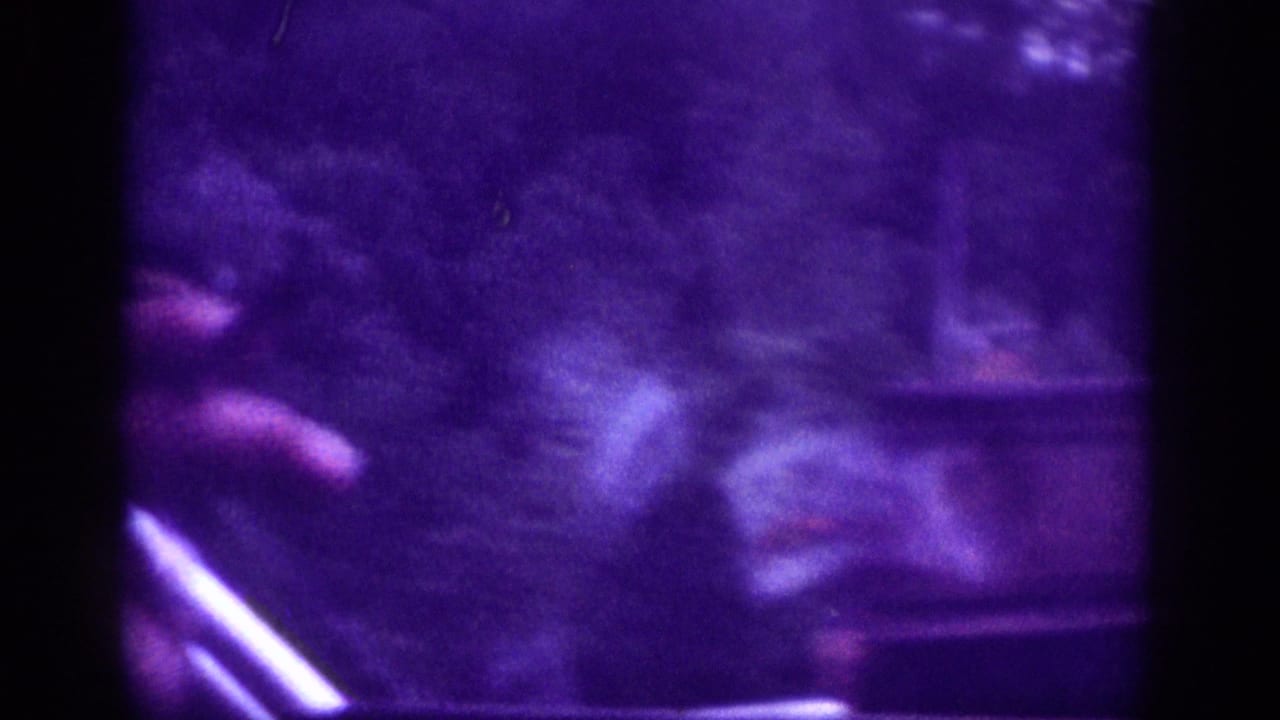 Blurred 8mm home-movie frame with a visible hand holding a small sign that appears to reference a catch or pay fishing offer, with indistinct roadside surroundings behind it.