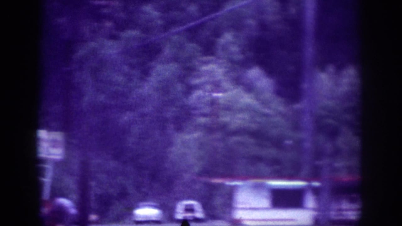 Purple-tinted 8mm frame of a suburban roadside with utility poles, a white station wagon, and dense tree canopy viewed from a moving car.