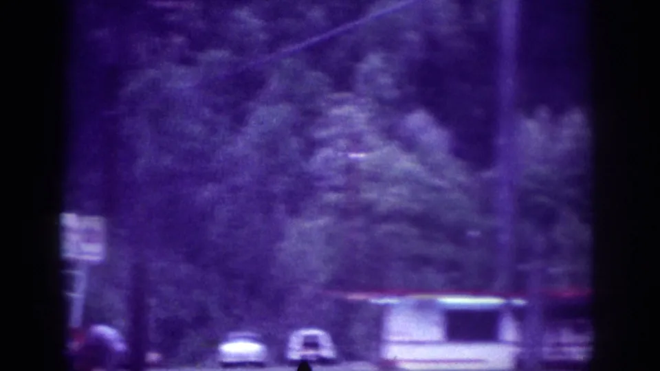 Drive-by 8mm view of a Memphis suburban roadside, circa 1963, with a white station wagon, utility poles, a small roadside figure, and heavy tree cover.