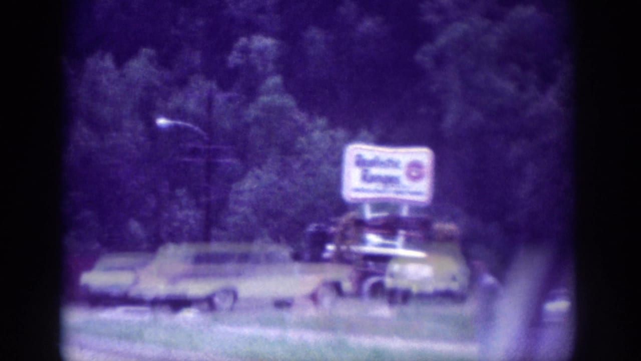Drive-by 8mm shot from a moving vehicle in Memphis, 1963, showing a yellow sedan beside a large roadside commercial signboard and street lamp.