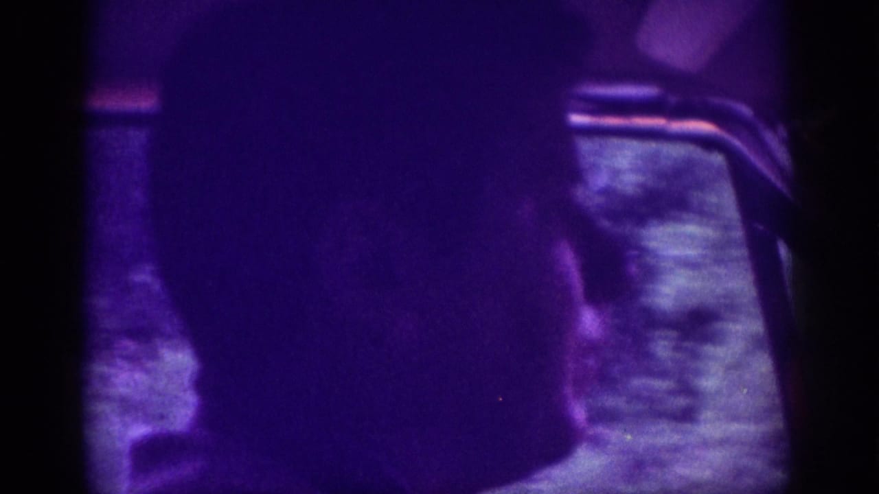 Silhouetted figure seen from behind against a car window interior dominated by a deep purple color cast.