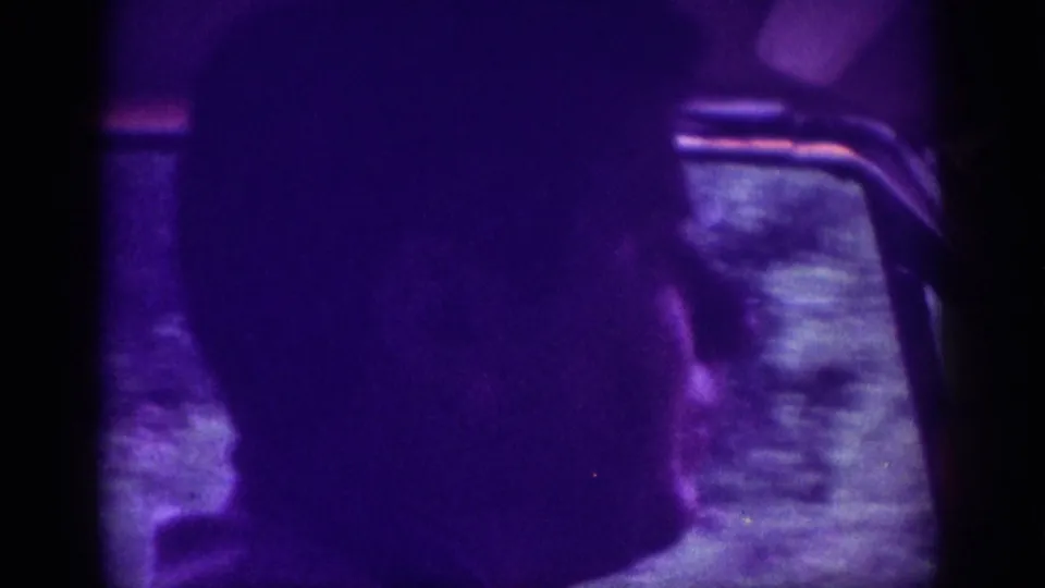 Silhouetted passenger profile against a car window, overwhelmed by a purple dye-shift; 8mm home-movie footage consistent with 1963.