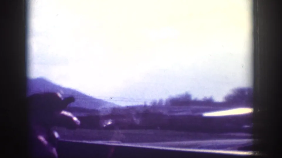 Through-windshield shot of a low rolling hill and tree line along a highway with overexposed sky; 8mm road-trip footage, circa 1963.