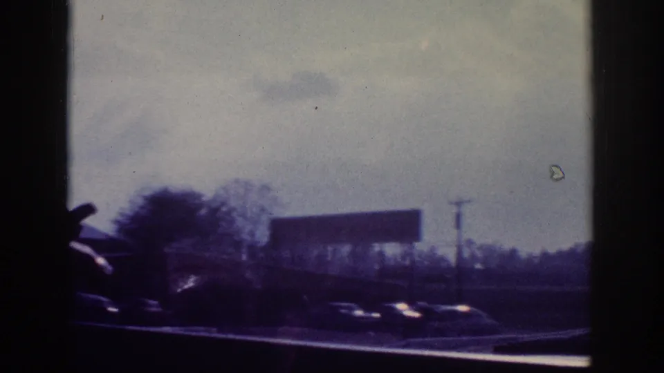Roadside highway view from inside a moving car, with a blank billboard and utility poles; 8mm footage consistent with early 1960s American highway travel.