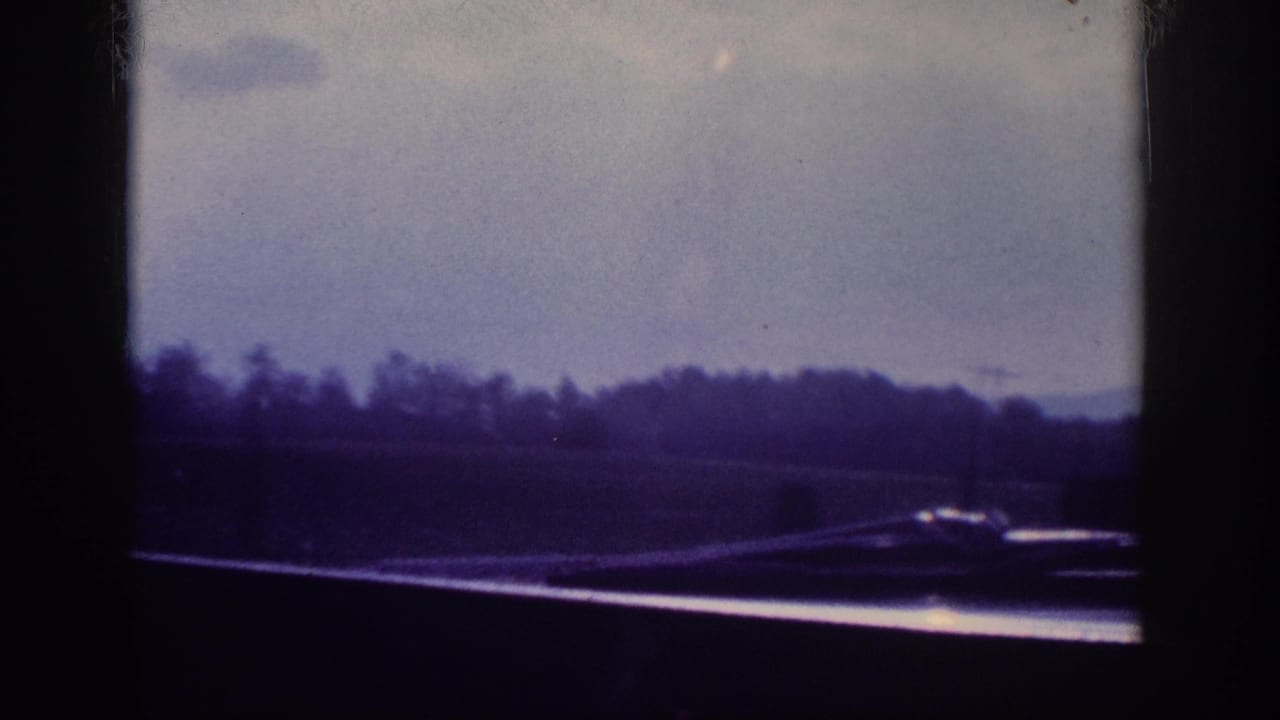 Highway road surface and treeline viewed through a windshield, heavily blue-shifted aged 8mm color film.