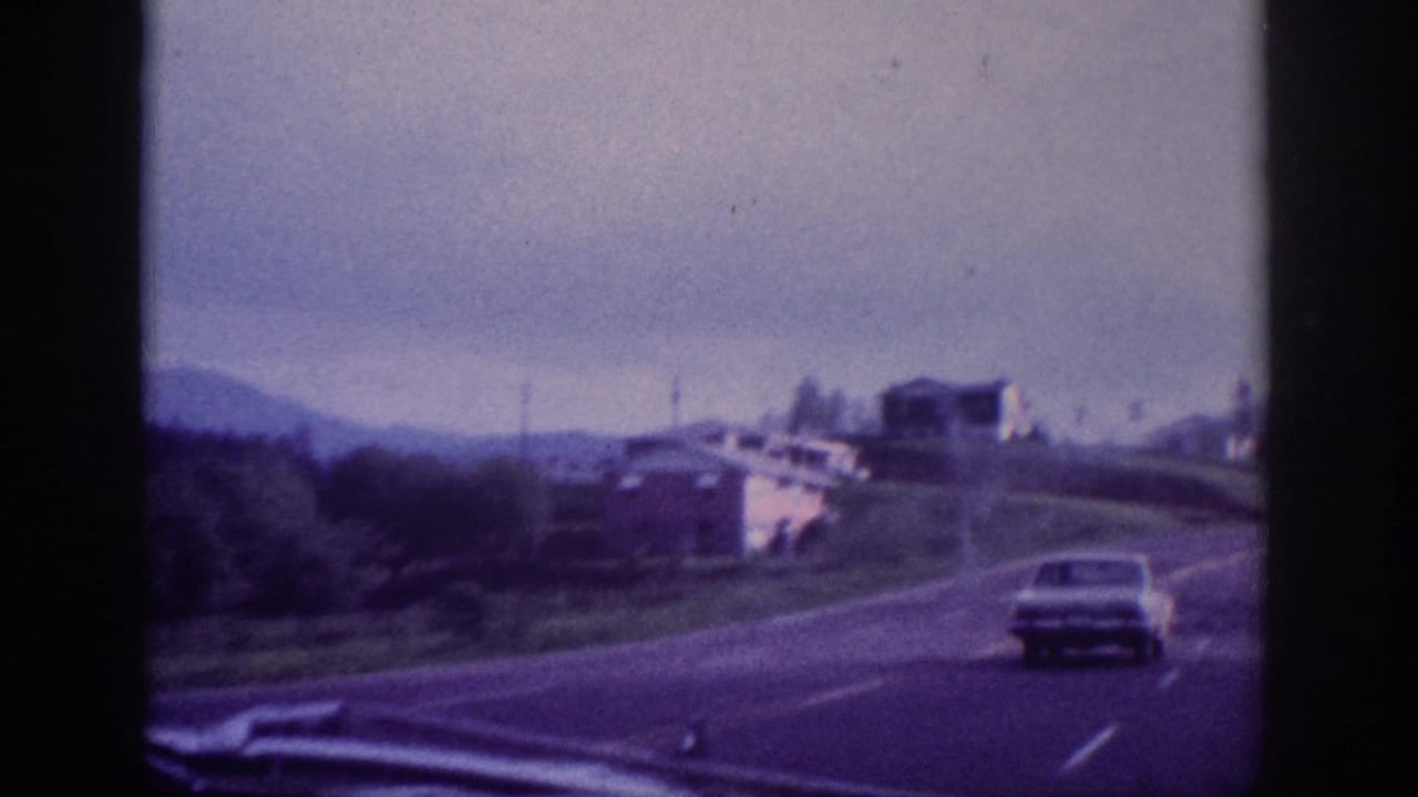 Light-colored early-1960s sedan on a divided US highway with a white building on a grassy embankment, through-windshield 8mm shot circa 1963.