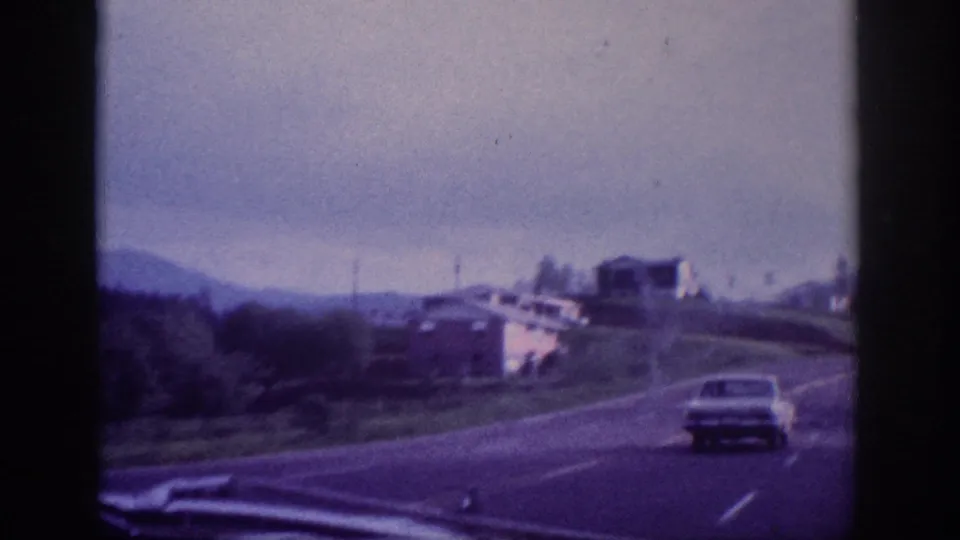 Light-colored early-1960s sedan on a divided US highway with a white building on a grassy embankment, through-windshield 8mm shot circa 1963.