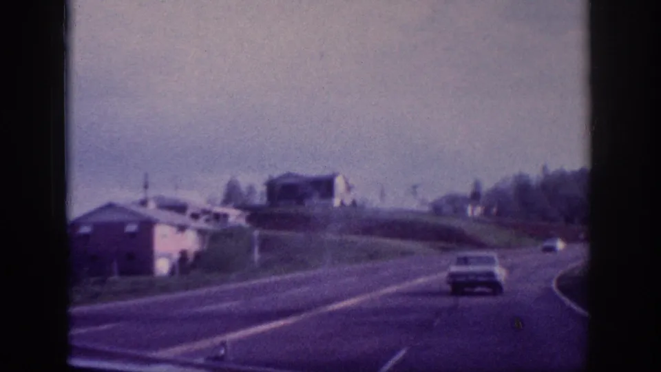 Sedan on a divided US highway with a low white building and grassy roadside embankment visible, 8mm color home movie attributed to Tennessee circa 1963.
