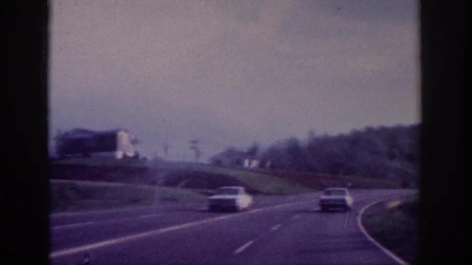 Two vehicles on a curving divided US highway; white roadside building and trees in background, 8mm home movie attributed to Tennessee circa 1963.