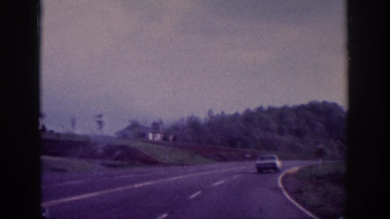 8mm home movie POV shot of a two-lane highway with a 1960s sedan visible ahead, tree-lined rolling hills under overcast sky, attributed to Tennessee area circa 1963.