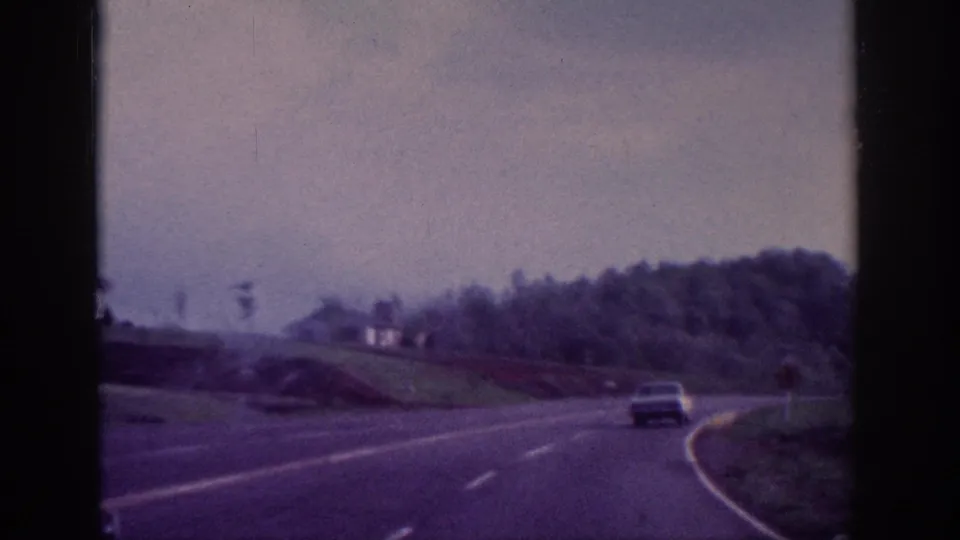 8mm home movie POV shot of a two-lane highway with a 1960s sedan visible ahead, tree-lined rolling hills under overcast sky, attributed to Tennessee area circa 1963.