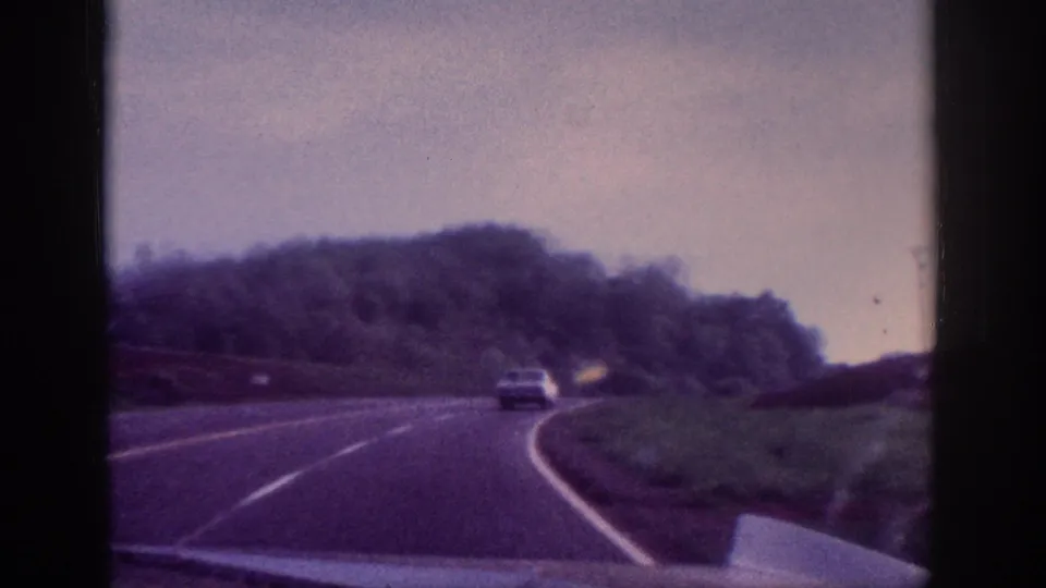 8mm POV driving footage showing a highway curve with a 1960s-era sedan ahead and tree-lined hills under grey sky, attributed to Tennessee circa 1963.