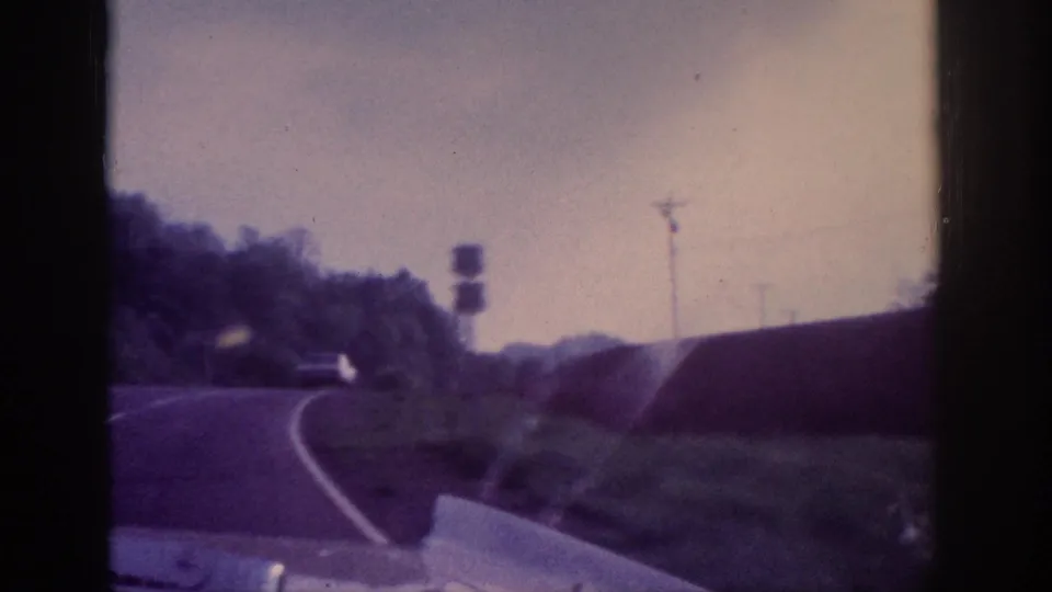 8mm POV frame showing a highway curve with an unreadable road warning sign and utility poles, pale washed-out sky and embankment typical of rural American road, early 1960s.