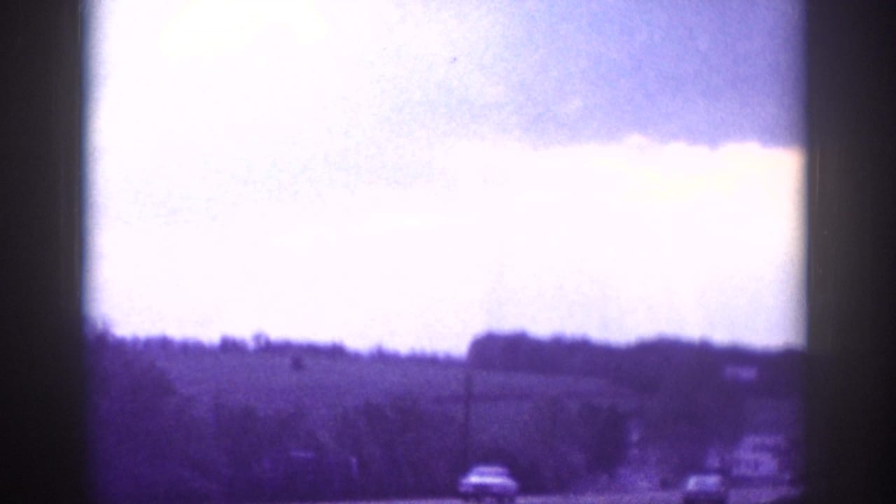 Overexposed purple-blue 8mm frame showing a suburban treeline, utility pole, and distant vehicles under a blown-out sky.