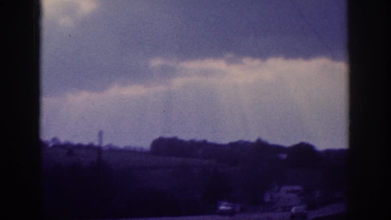 Purple-blue 8mm frame showing a dramatic cloudy sky, a distant tall structure on the horizon, and vehicles in the lower portion.