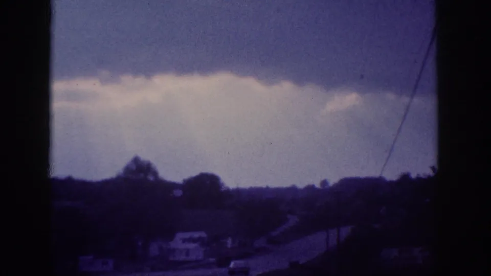 Blue-tinted 8mm frame showing a dramatic cumulus cloudbank above a suburban treeline, with a utility wire crossing diagonally, possibly from the Memphis outskirts circa 1963.