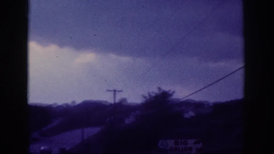 Closing 1963 8mm frame showing a utility pole, overhead power lines, and suburban rooftops under a cloudy sky; a possible attribution mark appears in the lower right corner.