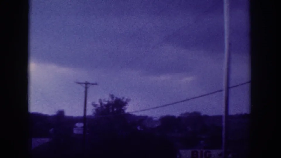 Aged 8mm home-movie frame from circa 1963 showing a stormy sky with a utility pole, power lines, and silhouetted rural treeline, shot through a car window.