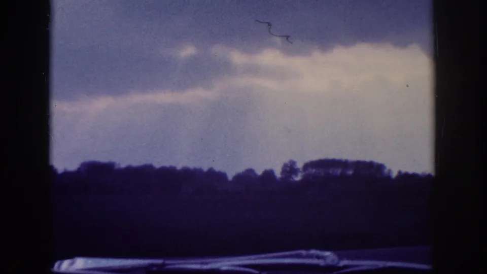 Aged 8mm home-movie frame circa 1963 showing dramatic cumulus clouds over a rural treeline, with a visible film-hair scratch artifact and a car edge at the frame bottom.