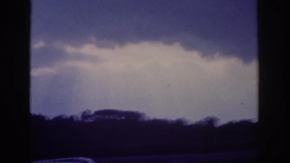 Aged 8mm home-movie frame circa 1963 showing a blue sky with cumulus clouds and a rural treeline, shot through a car window in the American South.