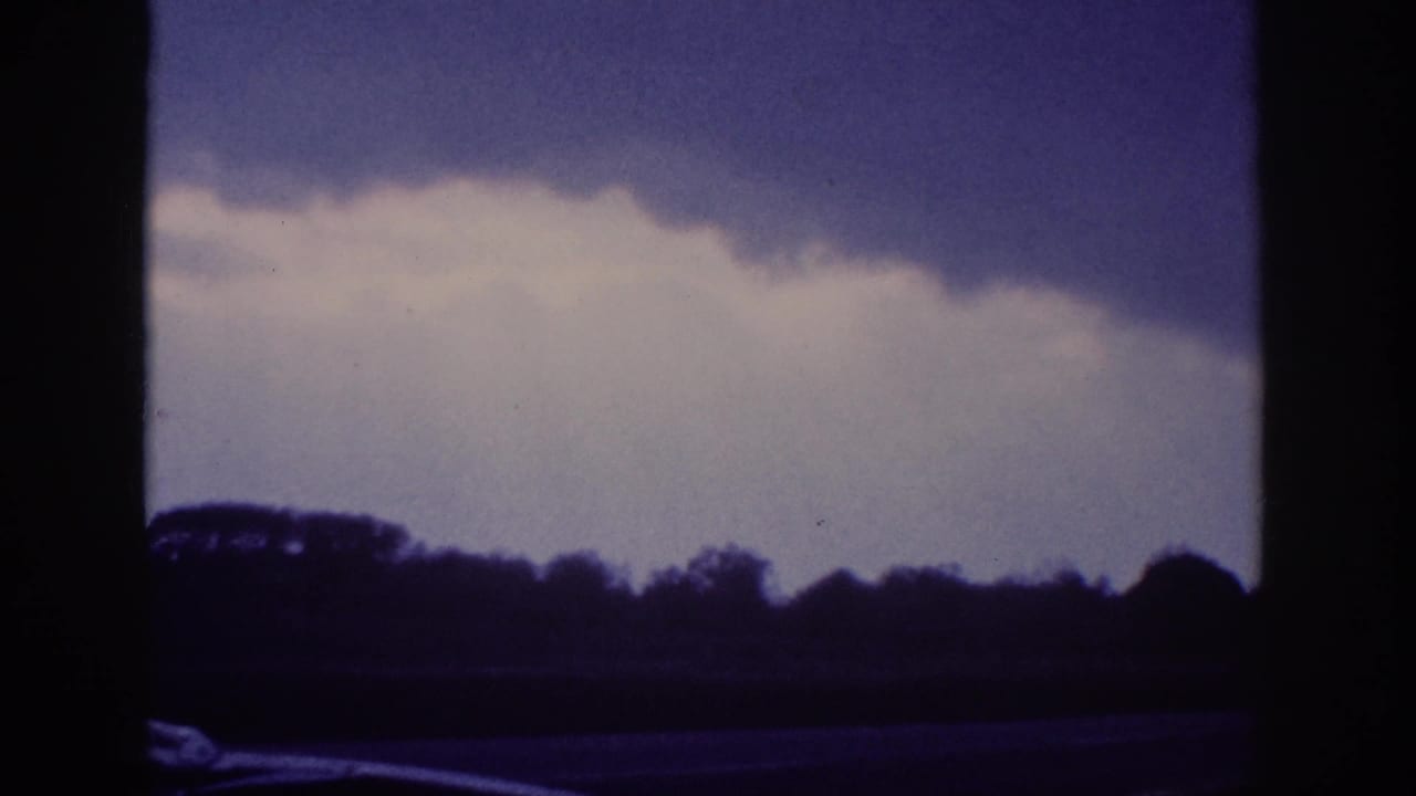 Stormy sky with billowing clouds over a silhouetted treeline, filmed from inside a moving vehicle, deep blue-purple 8mm film tint.