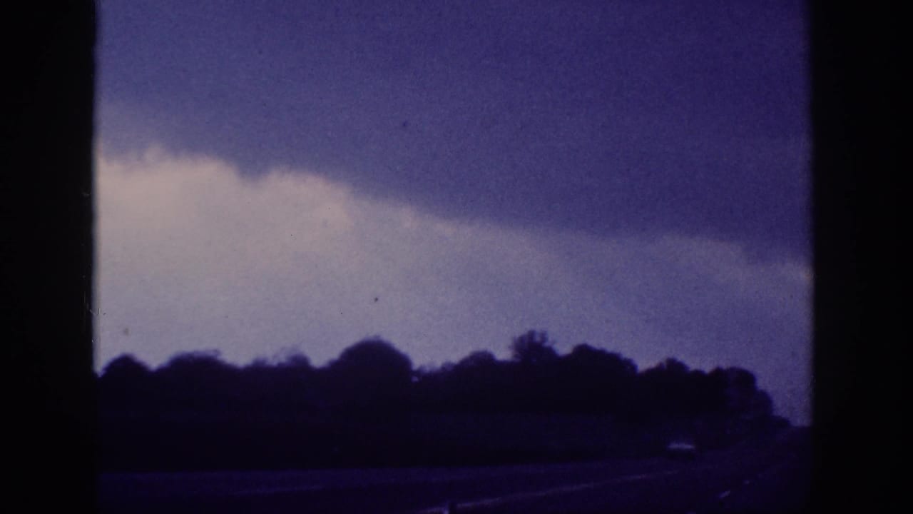 Storm cloud formation over dark treeline seen through car window, slightly shifted cloud arrangement, consistent purple-blue 8mm film cast.
