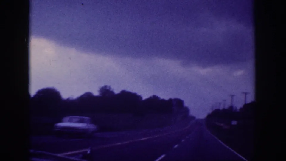 1963 8mm road footage — two-lane American highway with a period car and utility poles under a stormy sky, blue-purple dye shift from aged film.